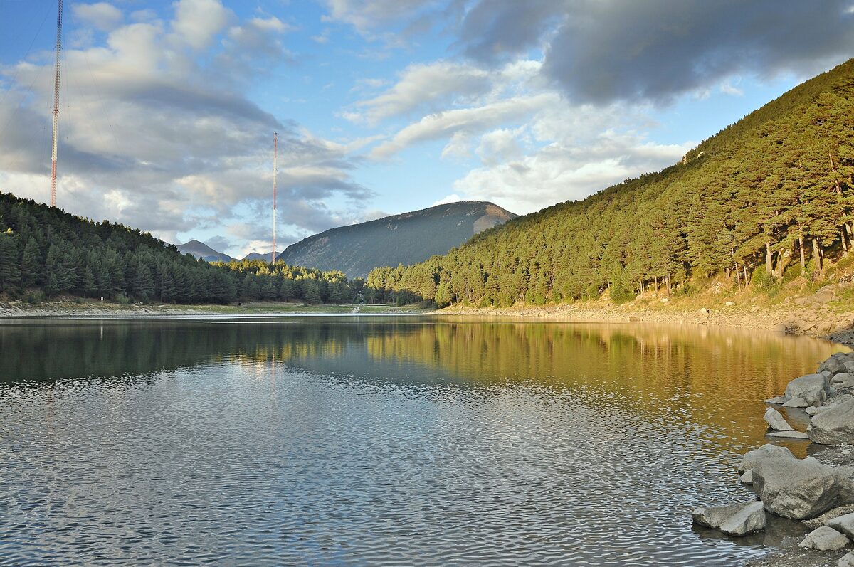 Lake Engolasters with a pine-covered hillside reflected in the still water and two radio masts behind the trees