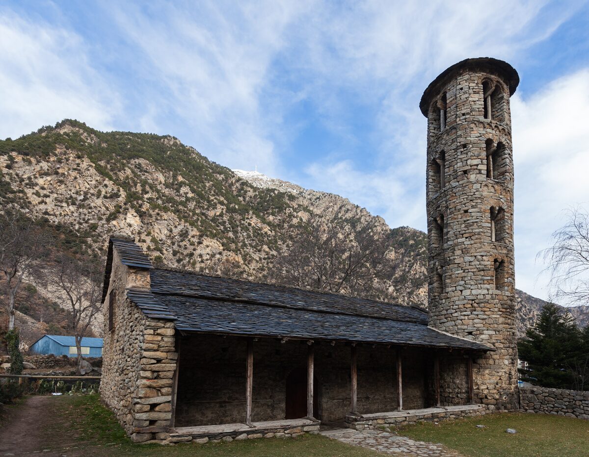 The Romanesque church of Santa Coloma with its tall round bell tower and slate-roofed porch, under a blue sky with clouds