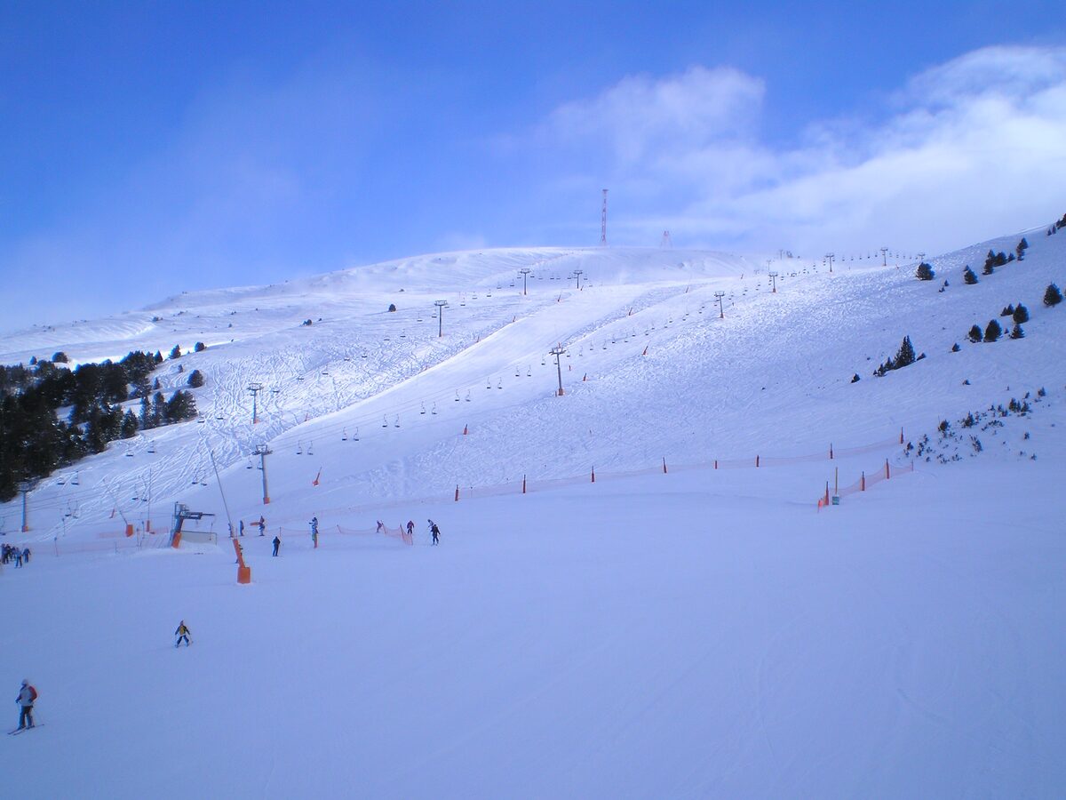 A wide groomed slope in the Grau Roig sector with a chairlift line climbing the hill and a few skiers behind orange safety netting
