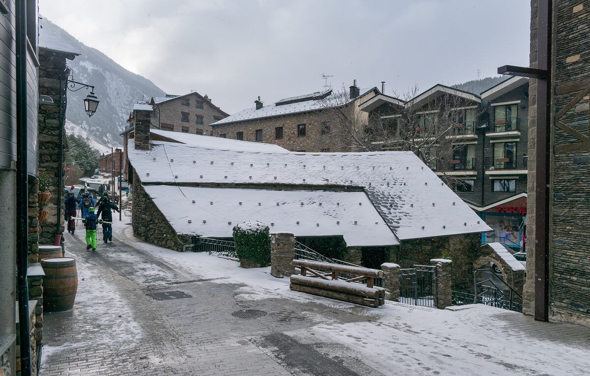 A snowy cobbled lane in Soldeu with skiers in colorful jackets walking past a slate-roofed stone building and timber lodges