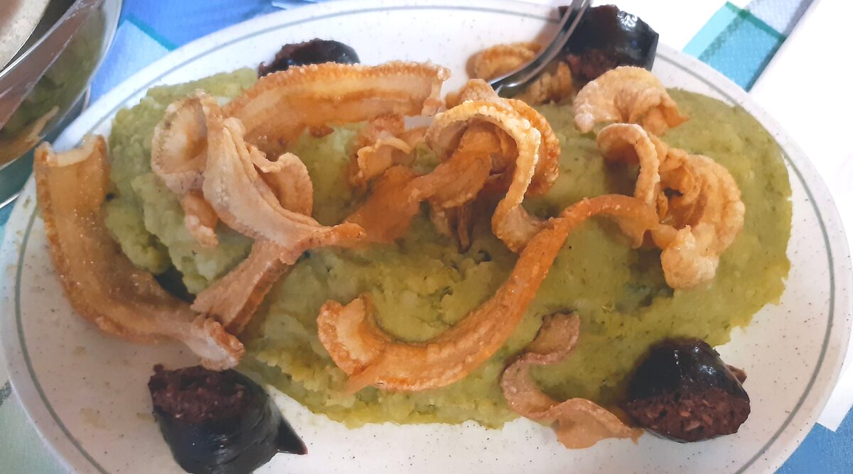 A plate of green mashed cabbage and potato topped with crisp fried pork rind strips, with slices of black pudding on the side