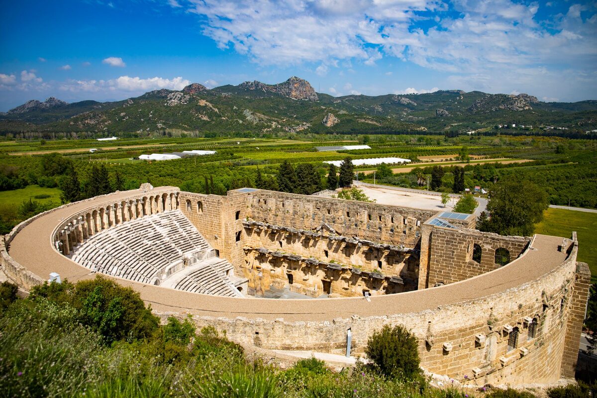 The Roman theater of Aspendos from the hill above, a full semicircle of seats and the stage building, with orchards and mountains behind