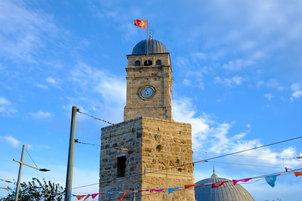 The stone clock tower of Kalekapısı with its lead dome and a Turkish flag against a blue sky