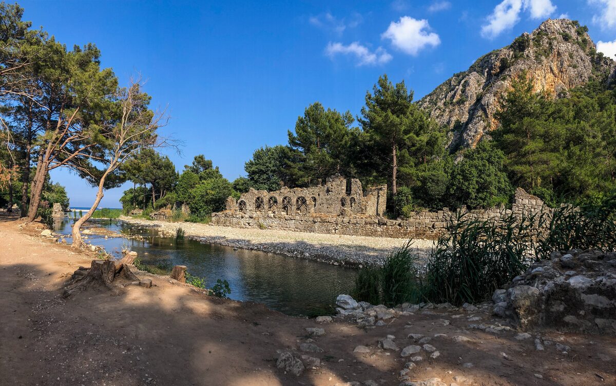 Arched stone ruins of Olympos by the stream among pines, under a rocky cliff