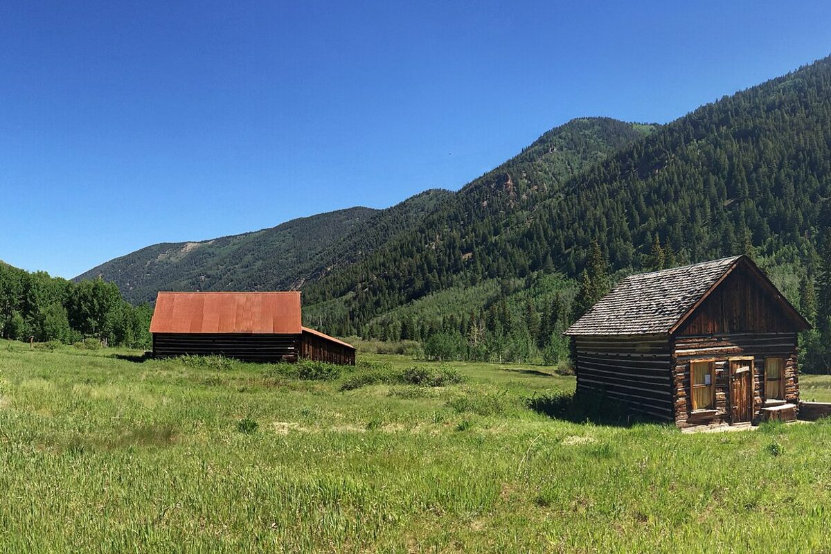 A rust-roofed barn and a weathered log cabin in the green Castle Creek valley at Ashcroft ghost town, photographed in summer
