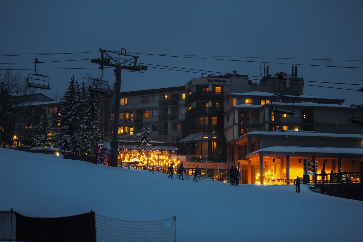 Snowmass Base Village at dusk in winter, with lit lodge windows, a chairlift tower and snow-covered slope in front