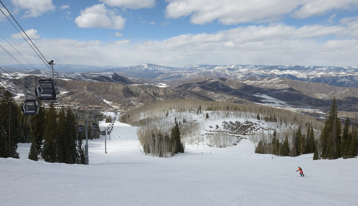 Gondola cabins above a snowy Snowmass run, with skiers on the slope and the mountains behind