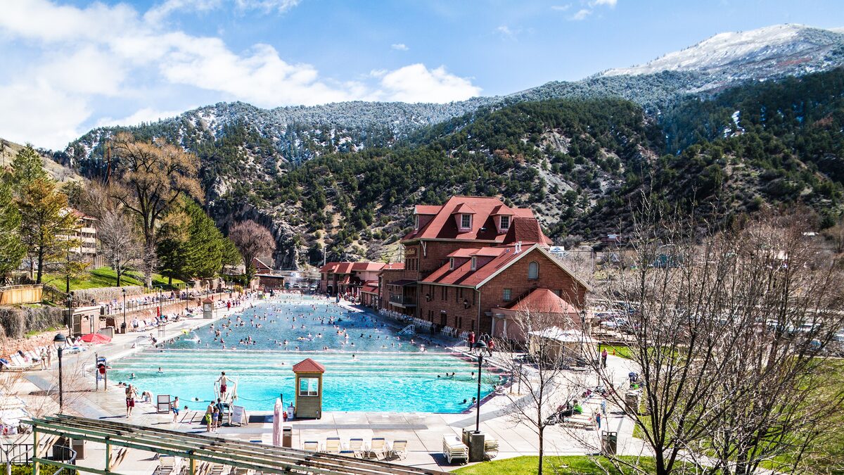 The big outdoor Glenwood Hot Springs Pool with swimmers, the red-roofed bathhouse and snow on the hills behind