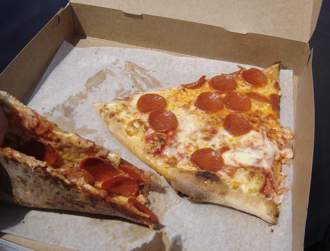 Two slices of thin-crust New York style pepperoni pizza in a takeaway box, one folded and partly eaten