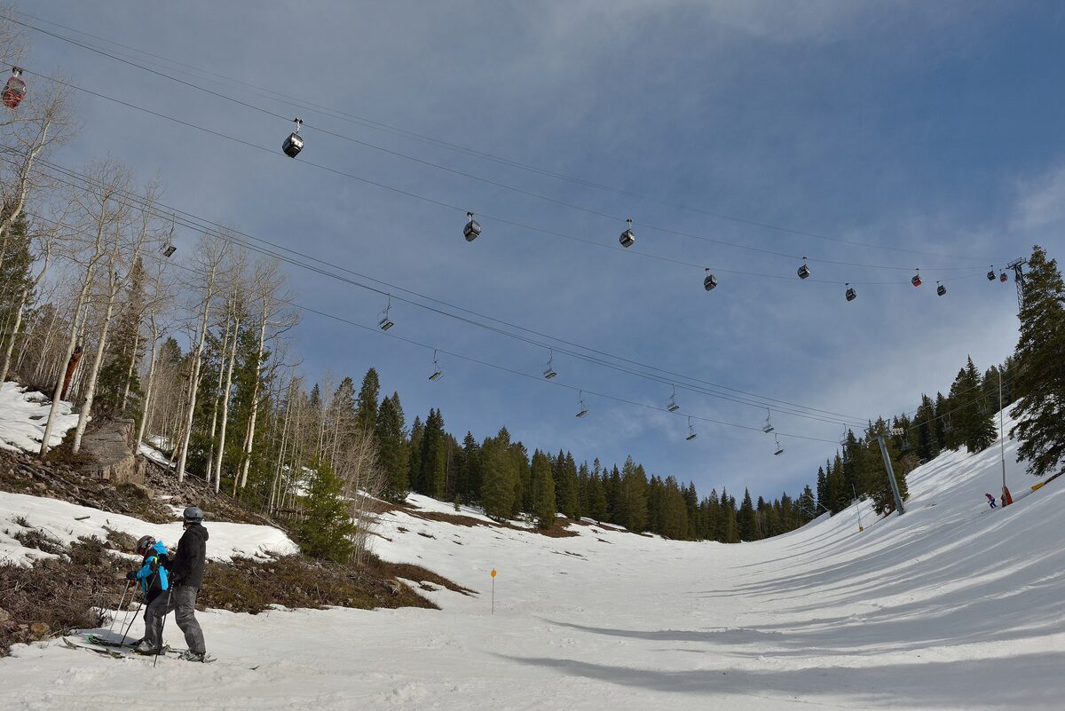 Gondola cabins over a snowy Aspen Mountain run, with two skiers and bare aspens beside the slope