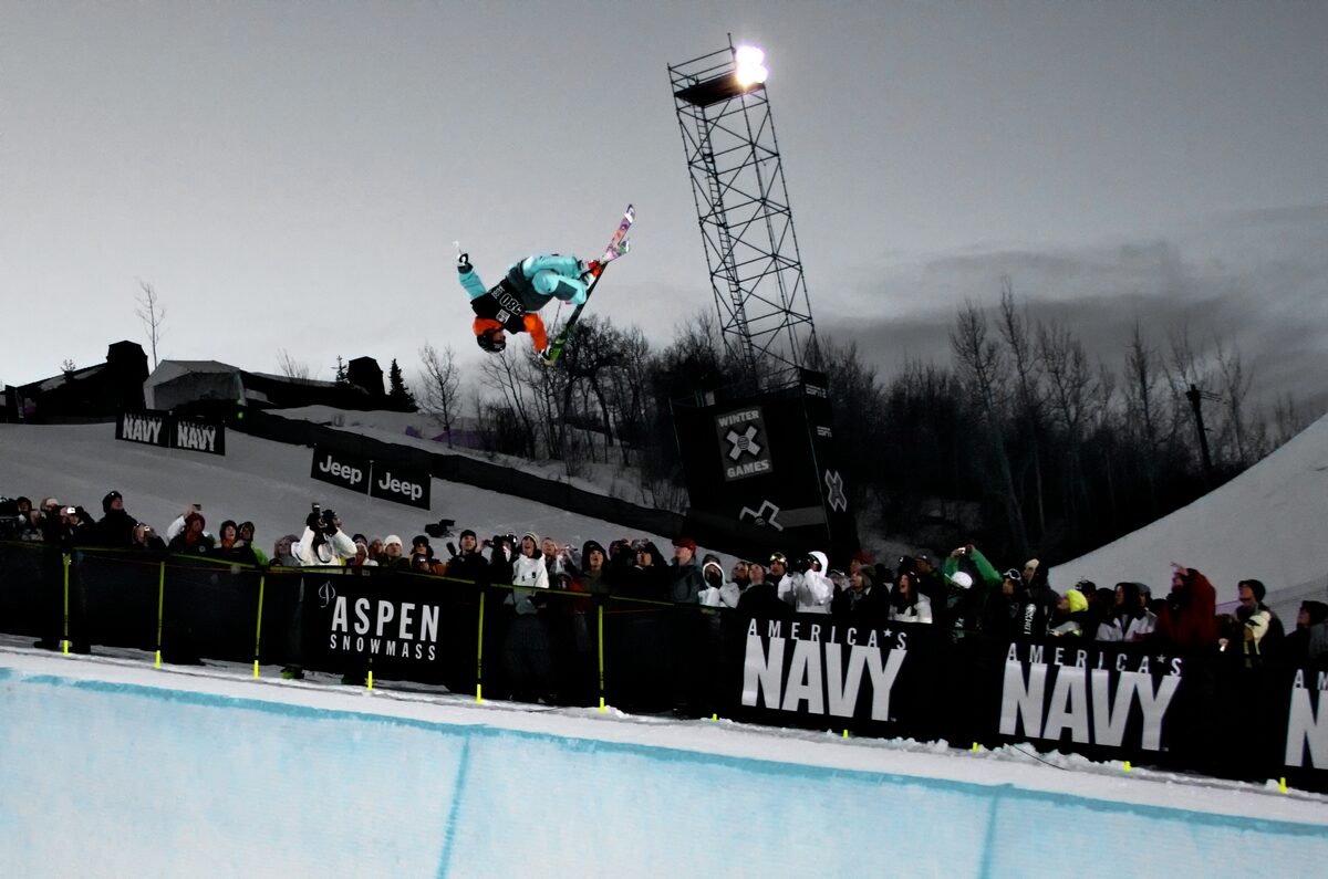A rider in mid-air at the Winter X Games in Aspen, with the Aspen Snowmass banner and the crowd behind the fence