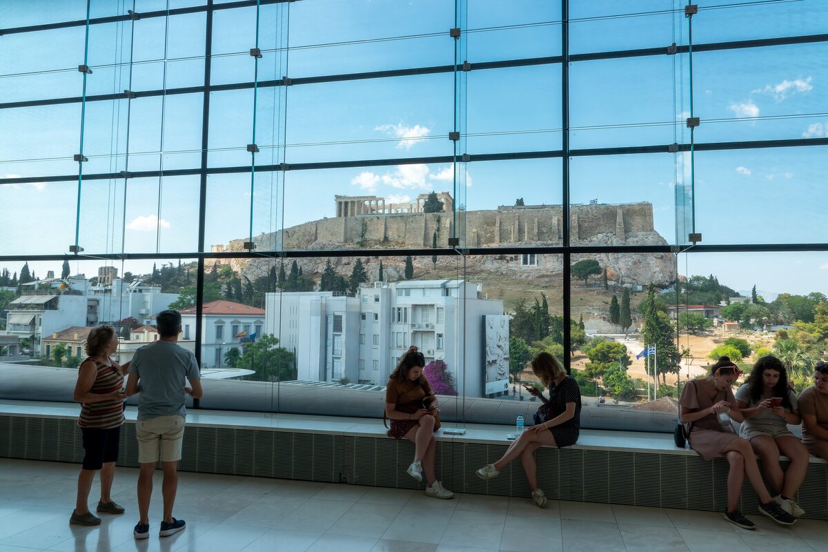 Visitors on a bench by the Acropolis Museum's glass wall, which frames the Acropolis across the rooftops