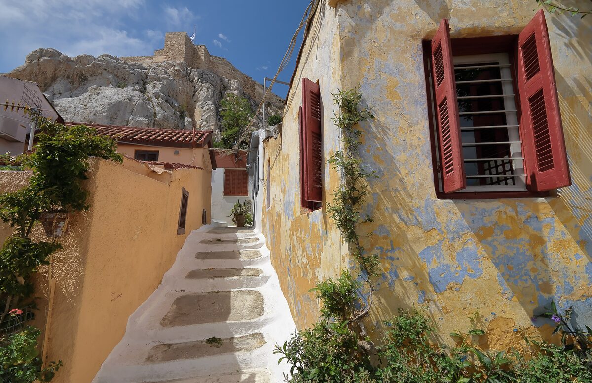 A narrow whitewashed stepped lane in Anafiotika with red shutters, under the rock of the Acropolis