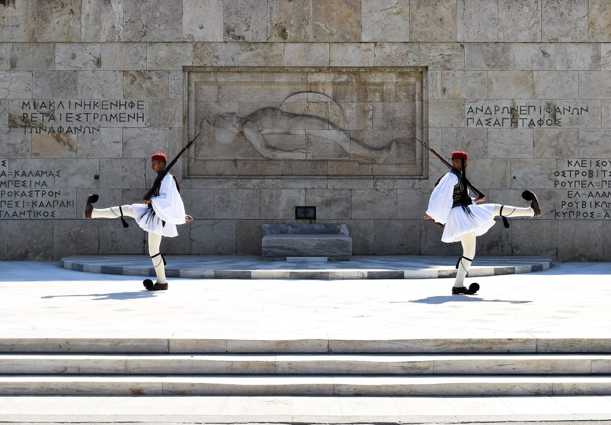 Two Evzones in white kilts and red caps, mid high-step, in front of the Tomb of the Unknown Soldier