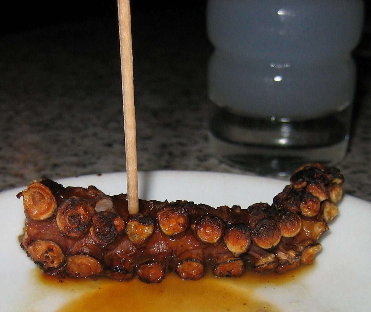 A piece of grilled octopus on a plate, with a glass of cloudy ouzo behind it