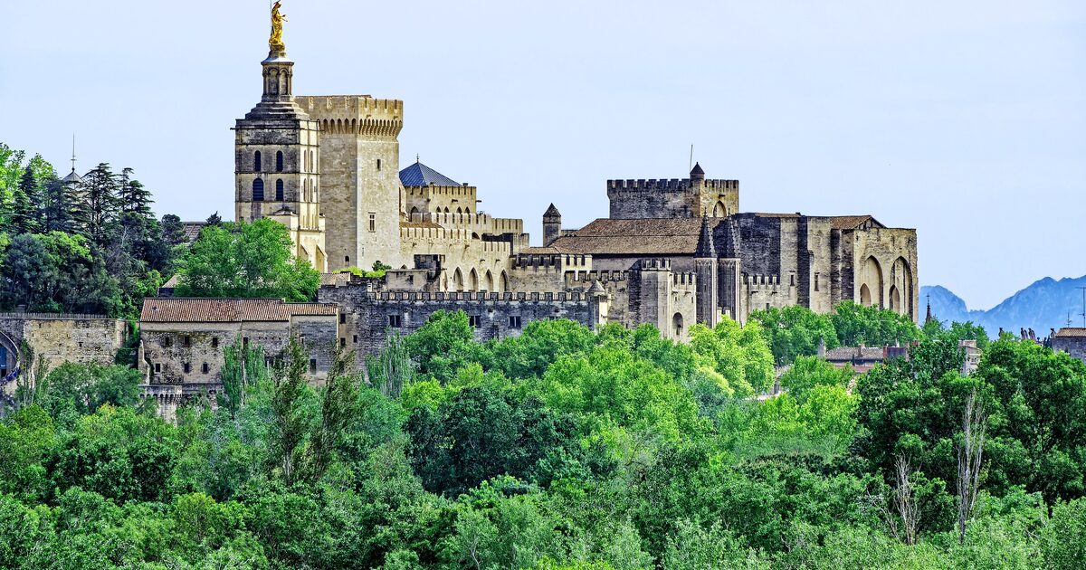 The Palais des Papes rising above green trees, with its towers and cream stone walls under a clear blue sky