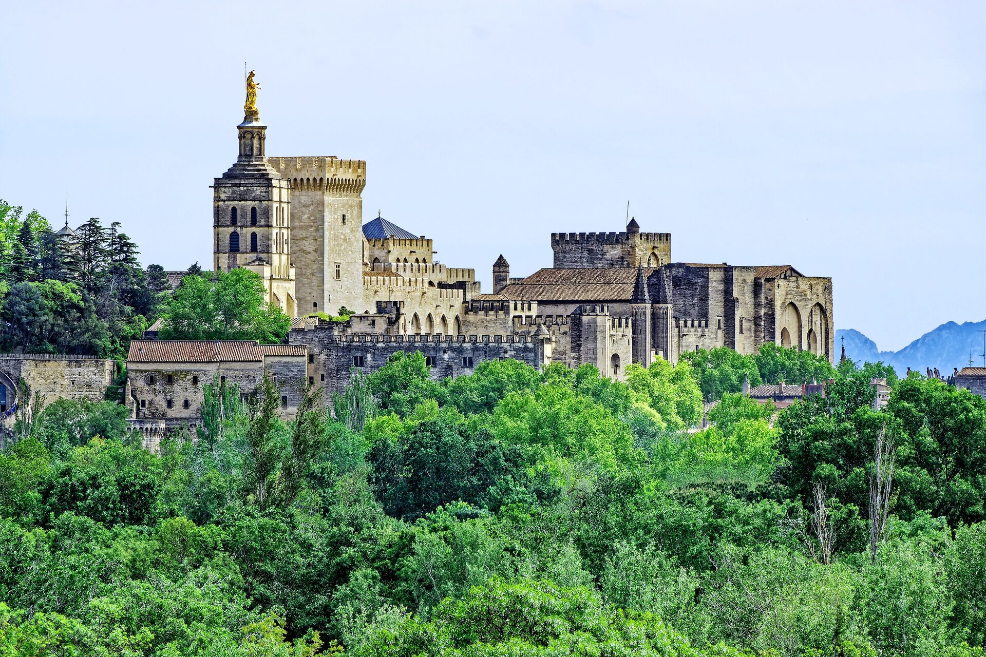 The Palais des Papes rising above green trees, with its towers and cream stone walls under a clear blue sky