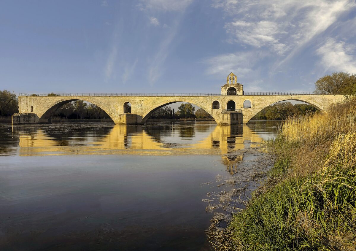 The stone arches of the Pont Saint-Bénézet over the Rhône, with the river reflecting the bridge in warm evening light