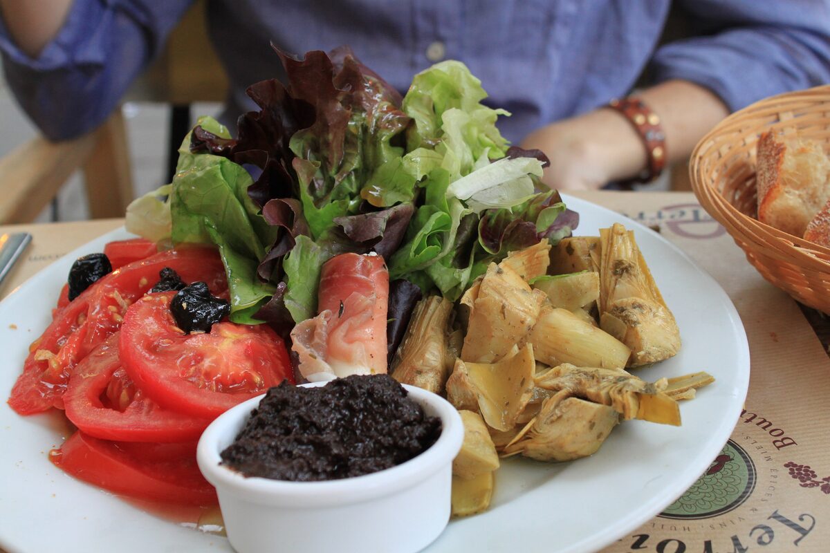 A salad plate with tomato slices, lettuce, artichokes and a small bowl of black tapenade