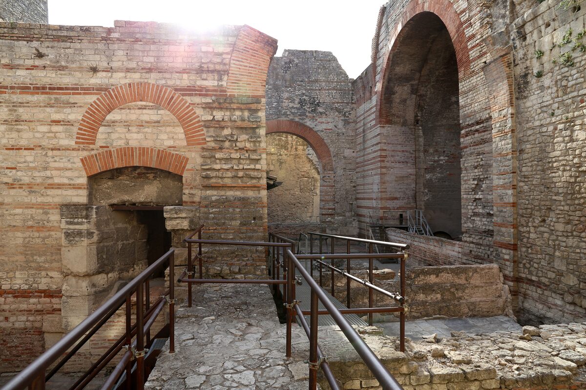 Brick arches and stone walls of the Baths of Constantine in Arles, with a railed walkway through the ruins