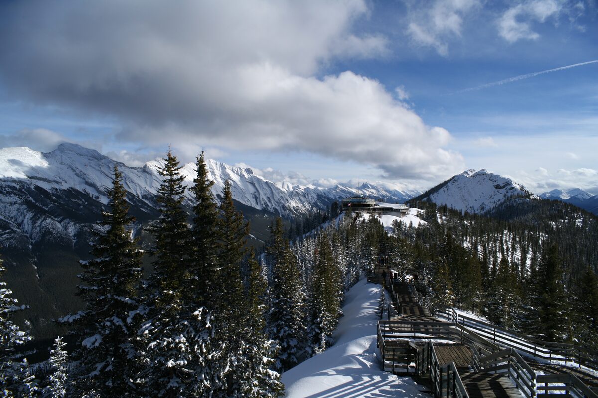 The summit boardwalk on Sulphur Mountain in winter, with snowy spruce, the upper gondola terminal on the ridge and mountain ranges beyond