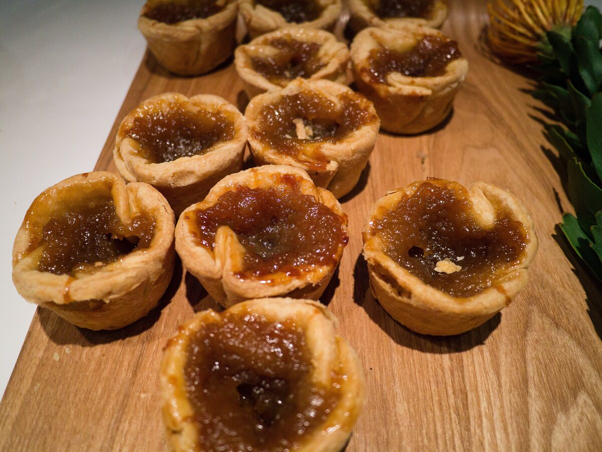 A wooden board of small butter tarts with golden pastry shells and a glossy, caramelized filling