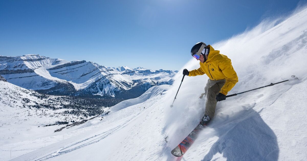 A skier in a yellow jacket carves through spraying powder at Lake Louise Ski Resort, with snow-covered peaks behind
