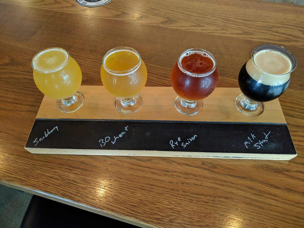 A tasting flight of four craft beers in small glasses, from pale gold to dark stout, on a wooden paddle
