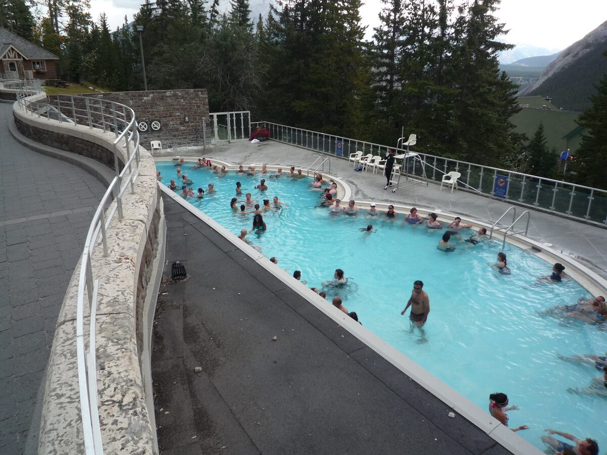 Bathers in the turquoise outdoor pool of the Banff Upper Hot Springs, with forest and a mountain behind (photographed without snow)