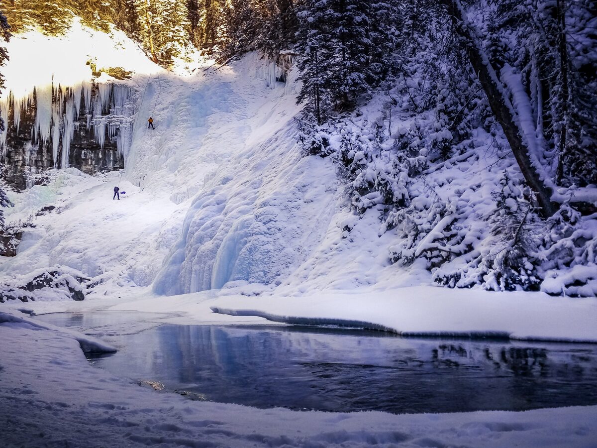 A frozen waterfall in Johnston Canyon, blue ice and snow-laden spruce with two tiny ice climbers on the face