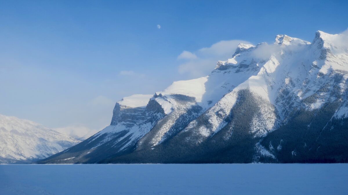 Frozen, snow-covered Lake Minnewanka in the foreground with a sunlit, snow-streaked mountain wall behind it
