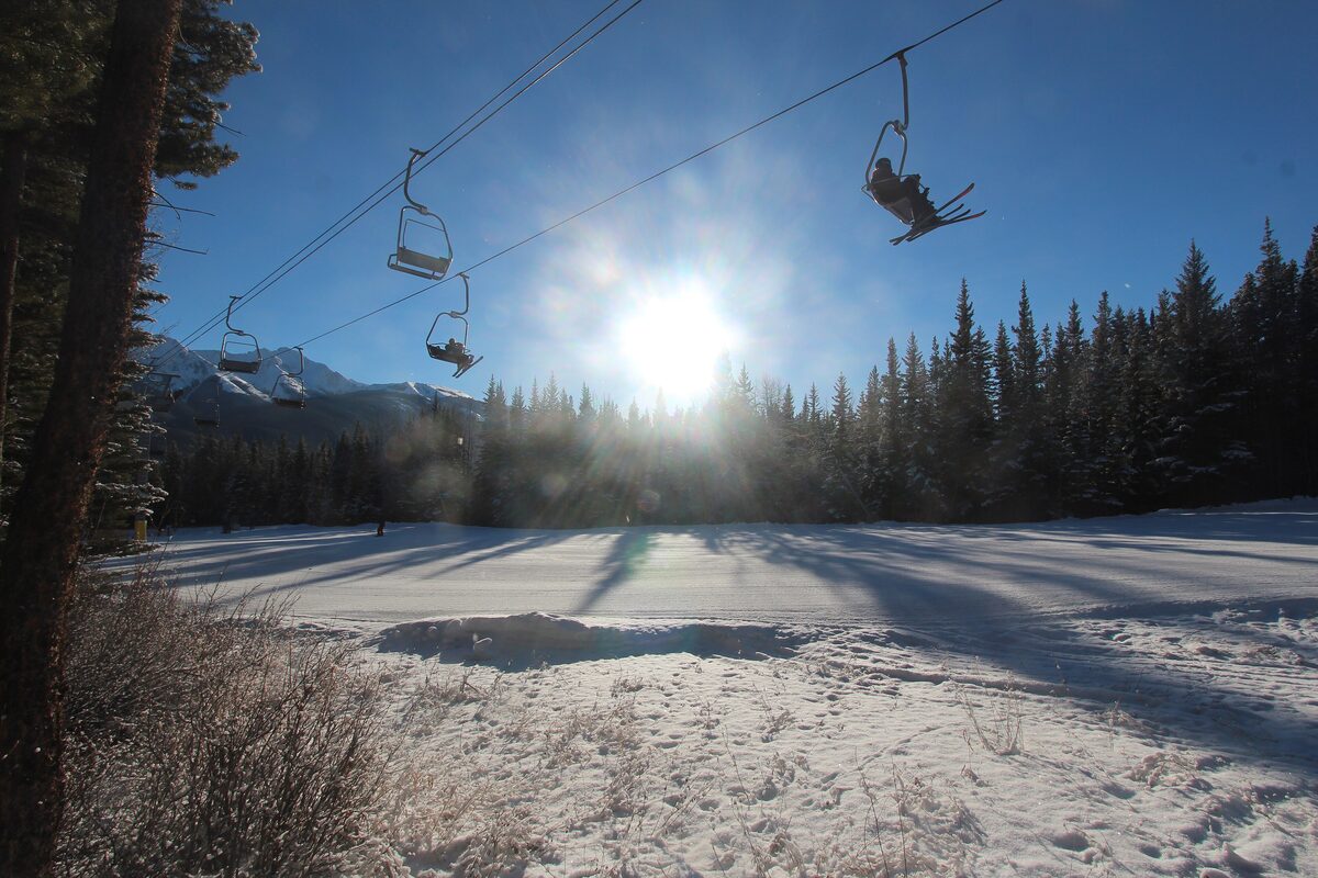 A chairlift carrying skiers above a snowy trail at Nakiska, with spruce forest and low winter sun