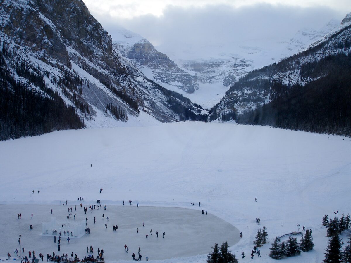 A crowd of skaters on the cleared rink on frozen Lake Louise, with snowy peaks and the glacier at the far end
