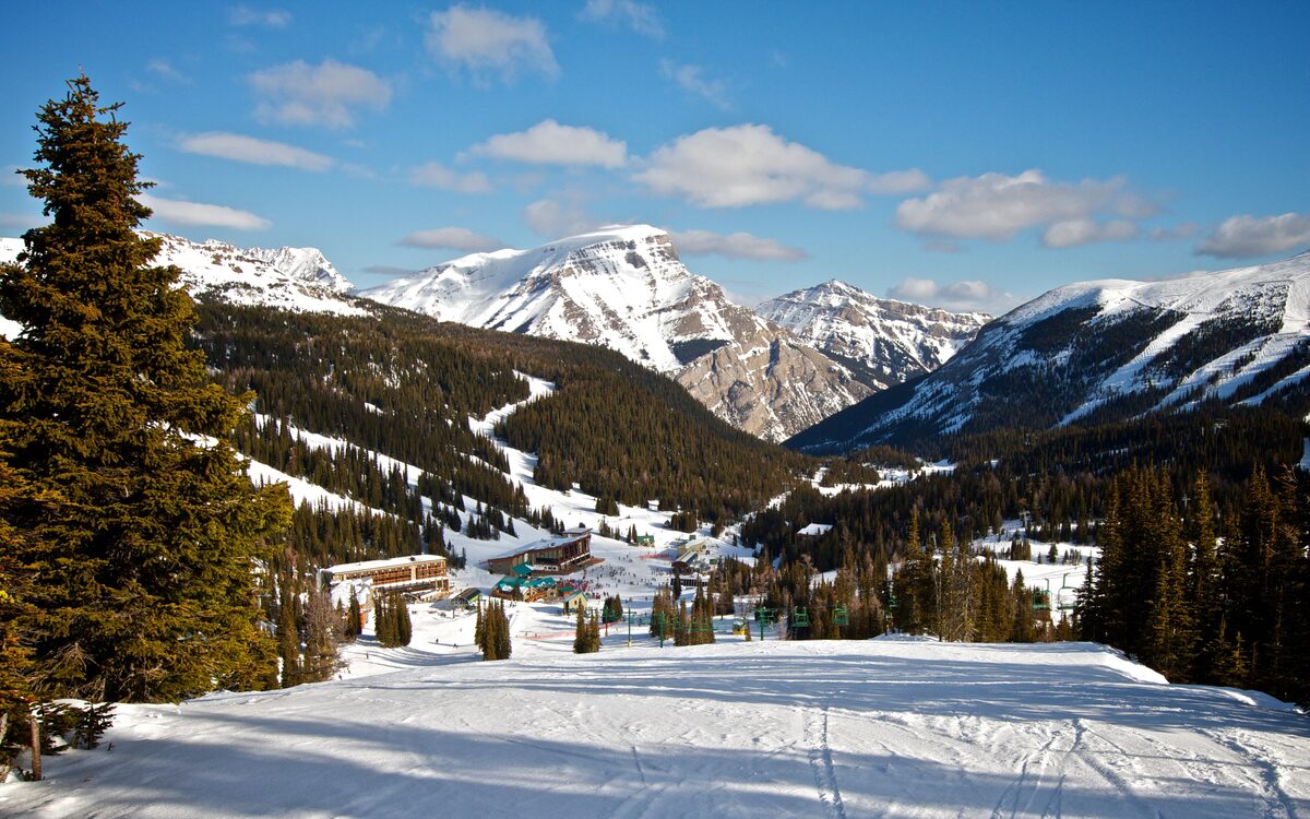 A groomed run leading down to the buildings of Sunshine Village, with snowy peaks and spruce trees around