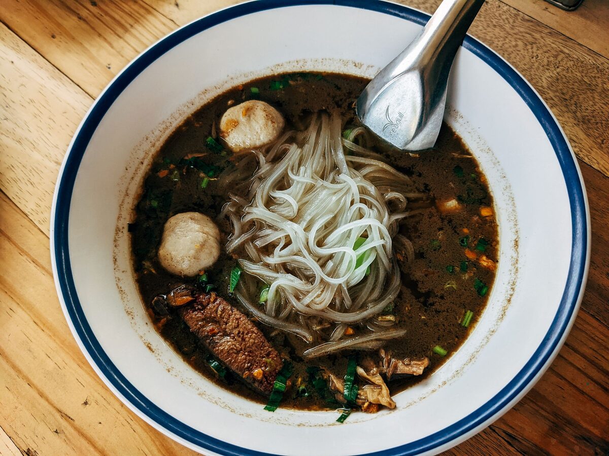 A white bowl with a blue rim holding dark-broth noodle soup with thin rice noodles and meatballs on a wooden table