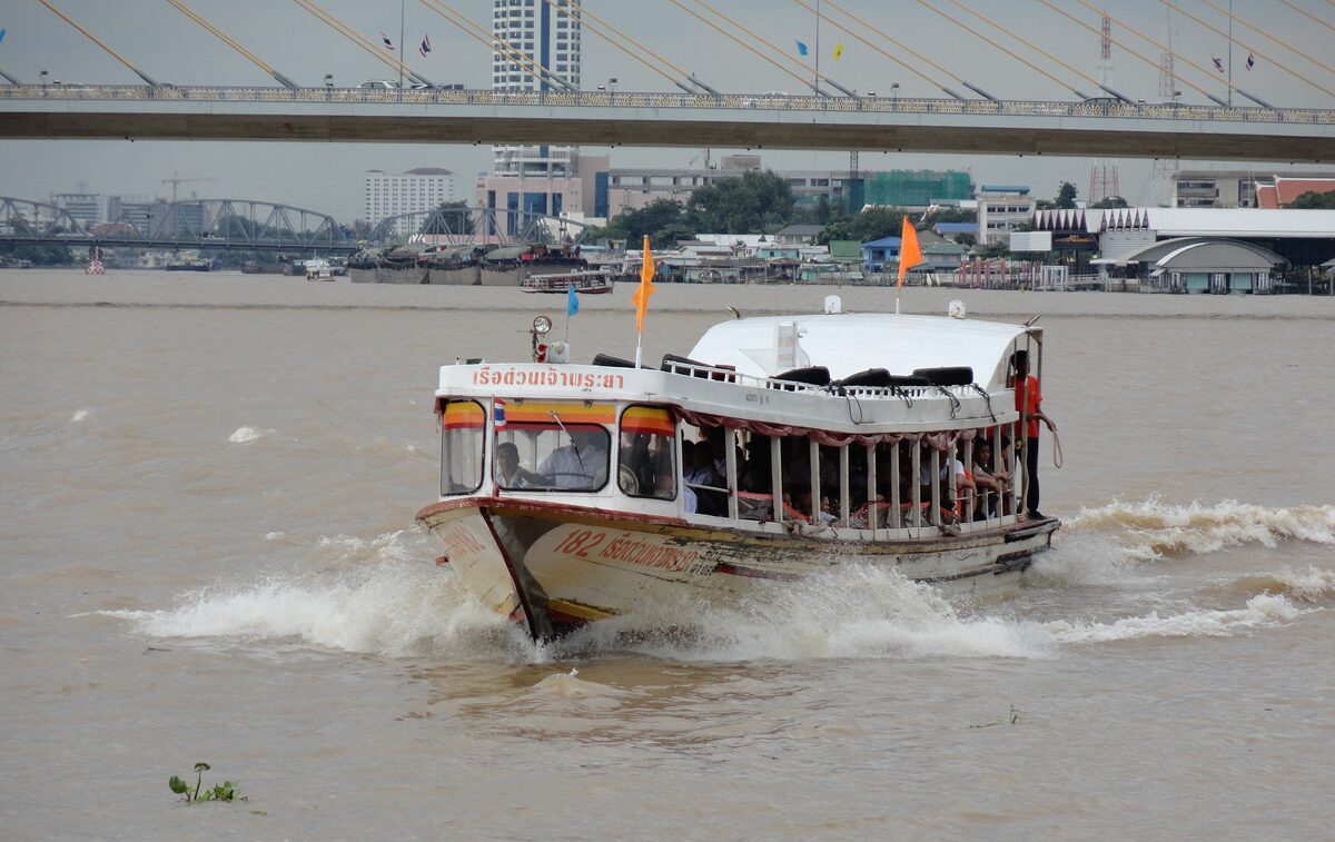 A passenger boat with a flag on its bow crosses the brown Chao Phraya River below a bridge