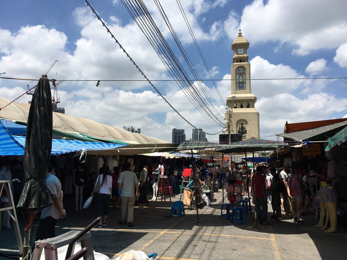 The clock tower of Chatuchak market rising above a crowded row of stalls under a blue sky