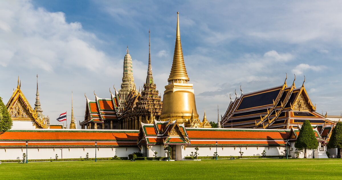 The golden prang and gilded halls of Wat Phra Kaew inside the Grand Palace, behind a long red-roofed wall and a Thai flag under a blue sky