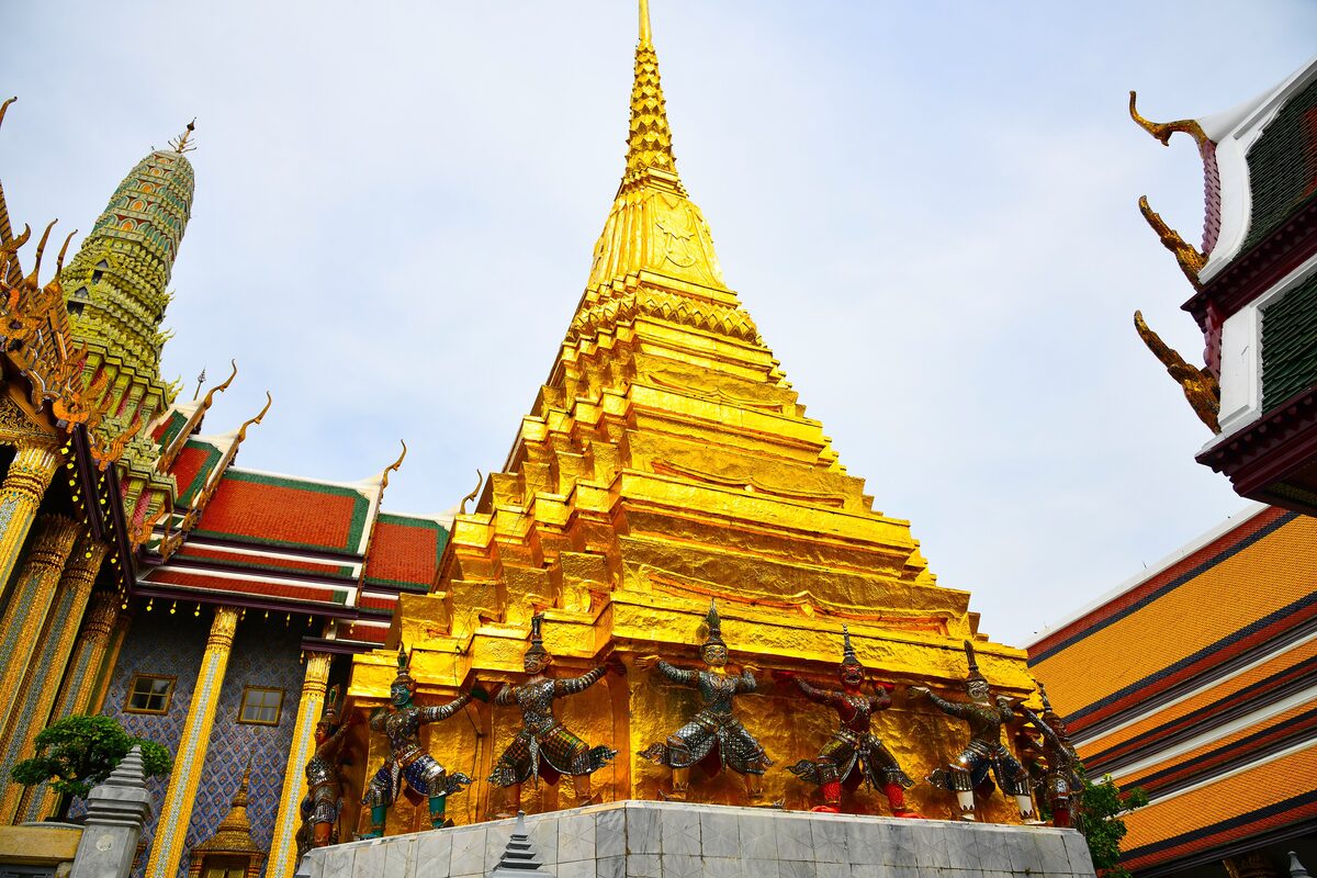 The golden chedi of Wat Phra Kaew with a row of demon guardian figures around its base, and red and gold temple roofs around it
