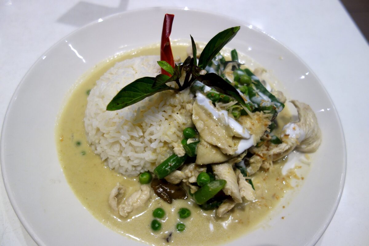 A plate of green curry with chicken, green beans and peas in a pale green coconut sauce, beside a mound of jasmine rice topped with Thai basil and a red chili