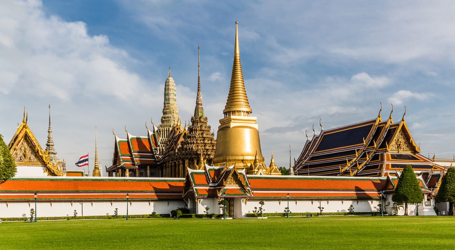 The golden prang and gilded halls of Wat Phra Kaew inside the Grand Palace, behind a long red-roofed wall and a Thai flag under a blue sky