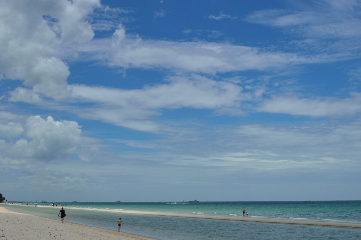 A long sandy beach at Hua Hin with a few people walking along the turquoise sea under a cloudy blue sky