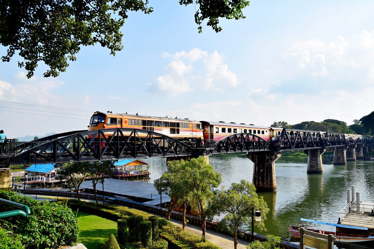 A passenger train crossing the steel truss River Kwai Bridge at Kanchanaburi on a sunny day