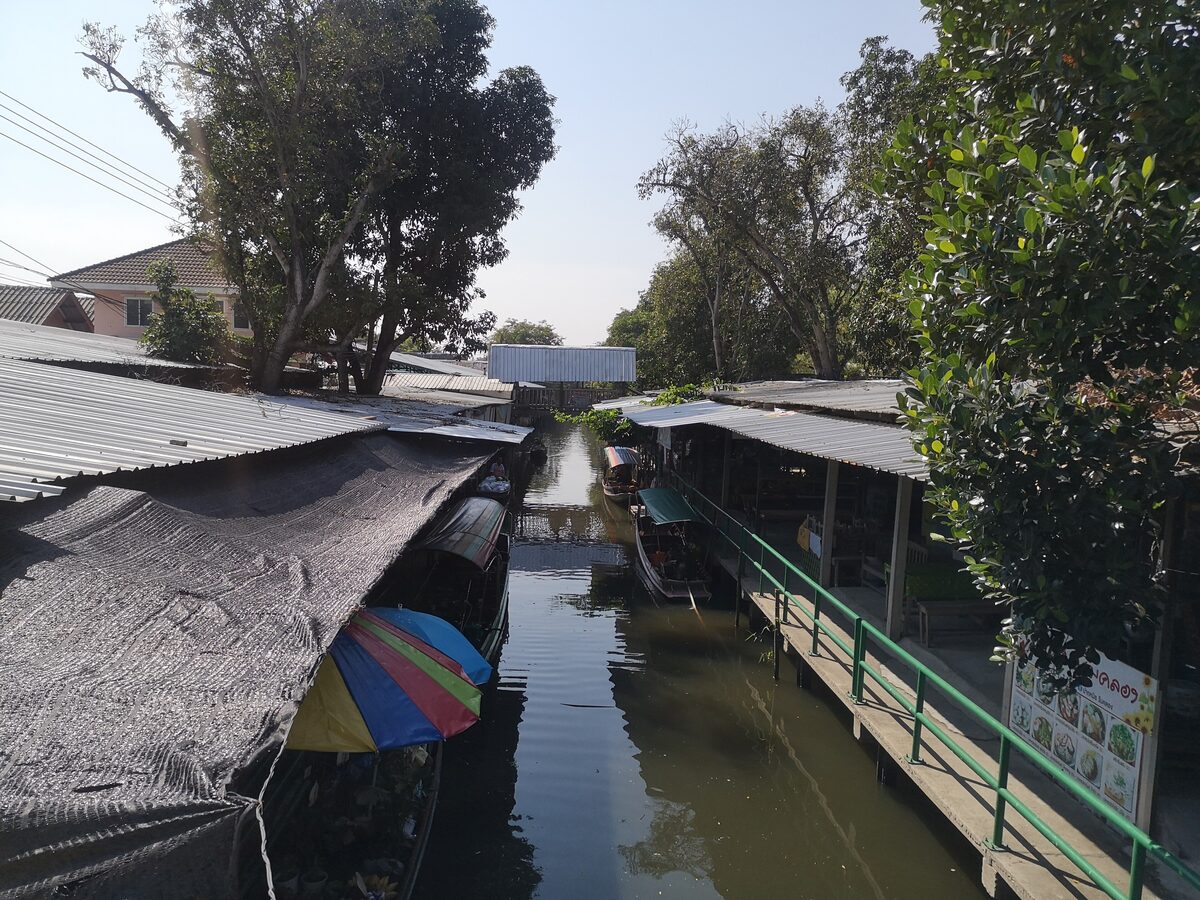 A narrow canal lined with tin-roofed market stalls and moored wooden boats, a colorful umbrella in the foreground, on a quiet day
