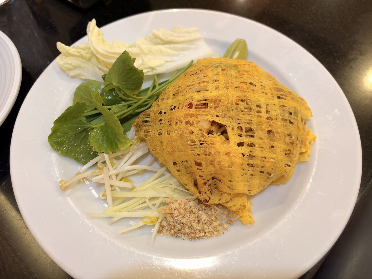 Pad thai wrapped in a lacy golden egg net on a white plate, with bean sprouts, banana blossom, herbs and ground peanuts beside it
