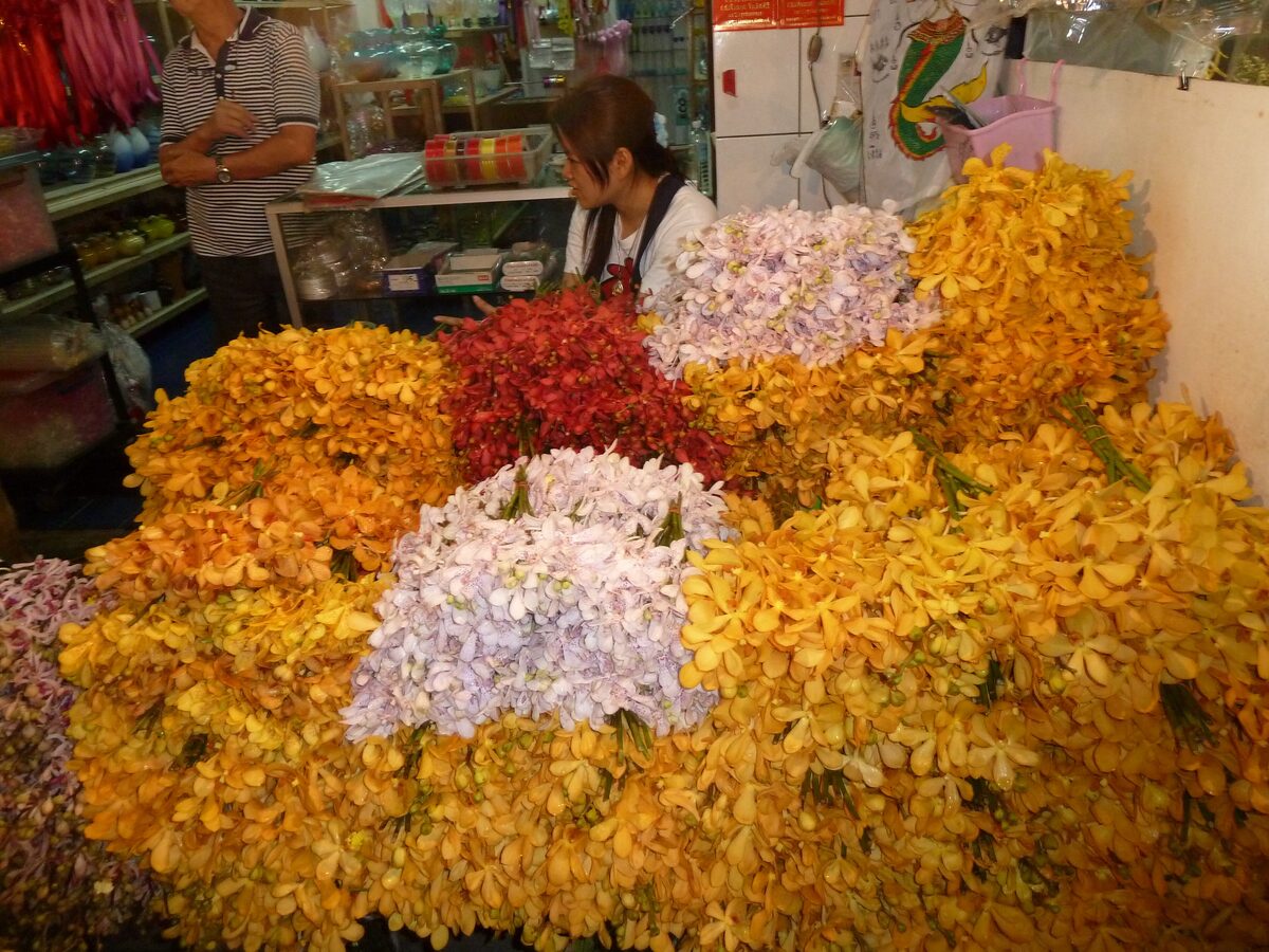 A flower stall at Pak Khlong Talat piled with yellow, red and pale lilac orchids, with two vendors small in the background