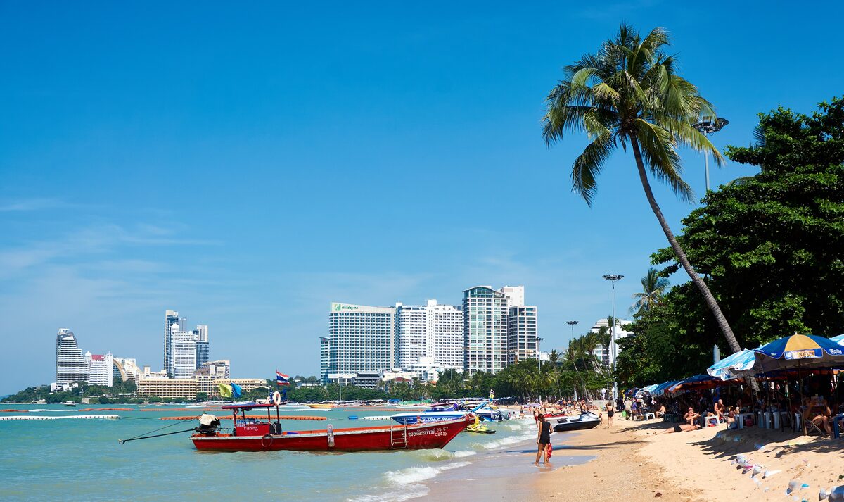 Pattaya Beach with hotel towers behind the sand, a red longtail boat at the shore, beach umbrellas and a leaning palm tree