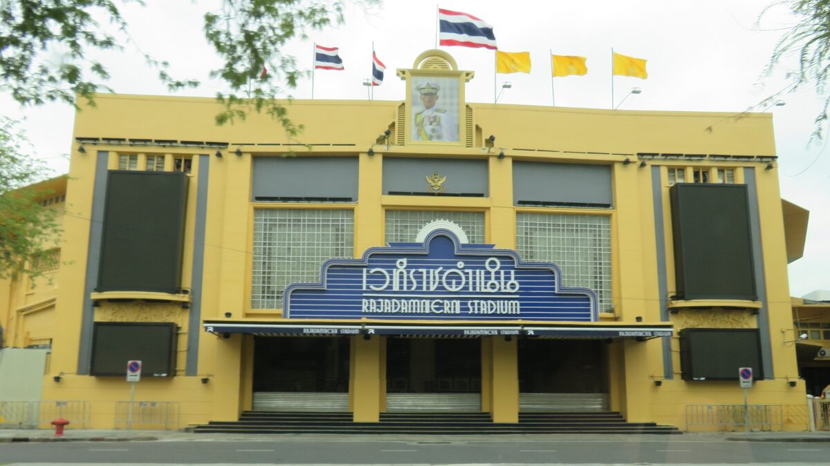 The yellow facade of Rajadamnern Stadium with its blue name sign above the closed shutters, under Thai and yellow royal flags