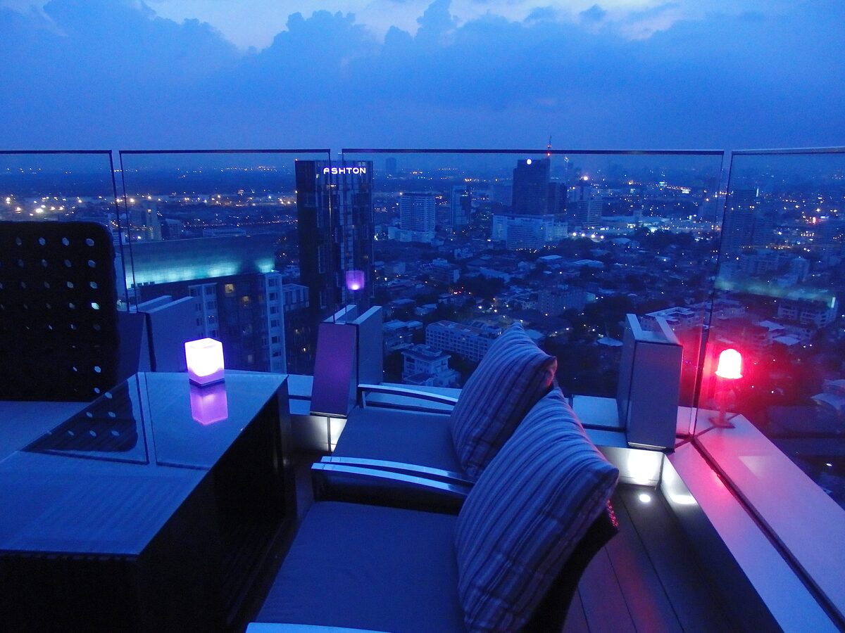Sofas and a low table on a high rooftop lounge in Bangkok at dusk, with glass railings and city lights spreading below