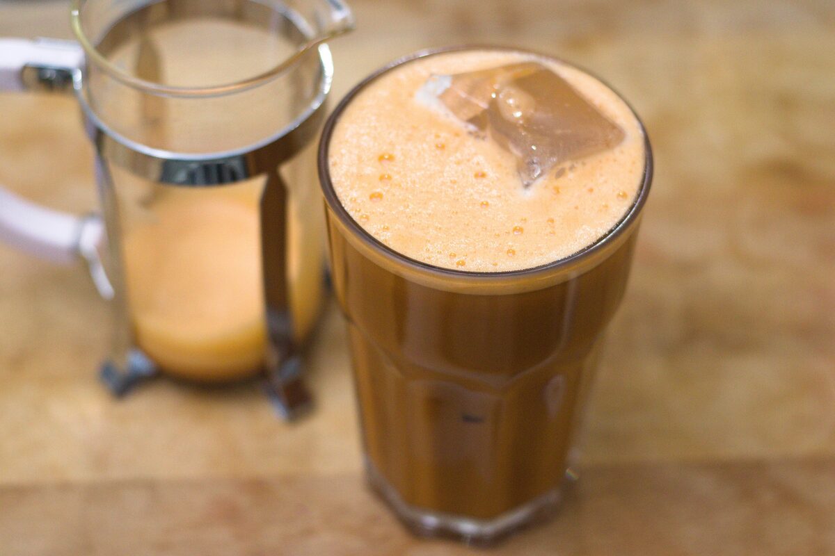 Two glasses of iced Thai milk tea, one foamy, beside a metal pitcher